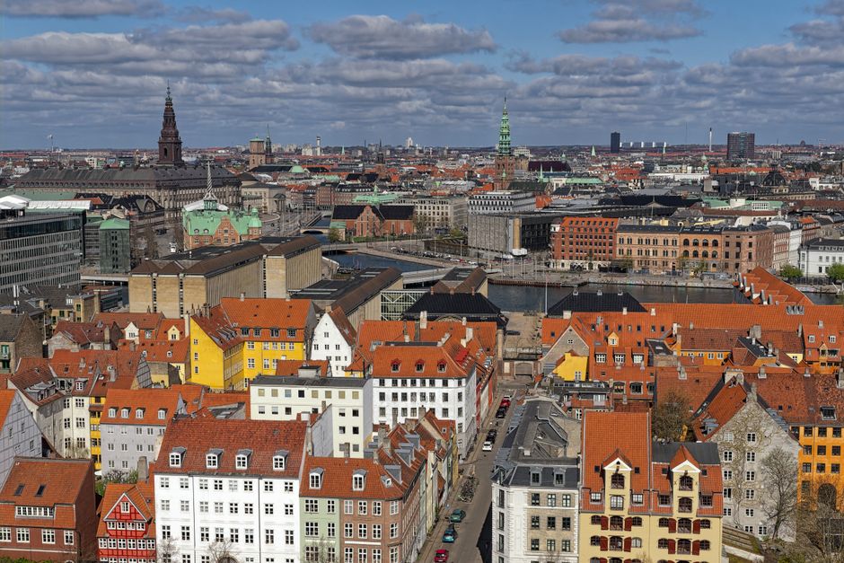 An aerial view capturing a cityscape with a large river running through the centre, surrounded by lush green parks and trees. Several bridges cross the river, connecting different parts of the city. In the foreground, residential houses with red-tiled roofs are visible among dense greenery. The middle ground features a variety of multi-storey buildings, including commercial and residential structures with flat and pitched roofs, arranged along busy streets with visible vehicular traffic. The background extends to a broad urban skyline under a partly cloudy sky, with some taller industrial or office buildings and a distant horizon. This scenery relates to home relocation and moving logistics, as seen in the context of professional removals services by Man with Van Denmark Hill, highlighting the transportation and packing environment involved in house removals in Denmark Hill.