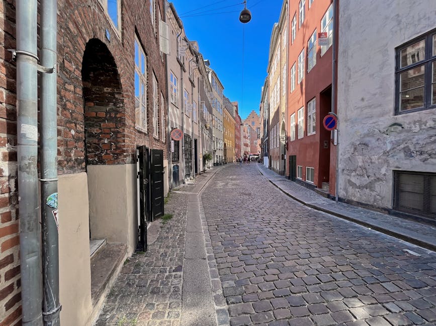 A narrow cobblestone street in Denmark Hill, lined with multi-storey buildings featuring a mix of brick and stucco facades in various colours, including beige, red, and yellow. The street is flanked by a concrete curb on the right and a paved sidewalk on the left, with black metal gates and doorway entrances visible along the buildings. Overhead, a single street lamp hangs from a wire suspended across the street, and two no-parking signs are mounted on the building walls. In the distance, there are a few pedestrians walking, and a few parked bicycles can be seen. The scene is illuminated by natural daylight, with shadows cast on the cobblestones, suggesting a clear sky. Although no moving vehicles or persons are directly visible loading or unloading, this setting reflects an urban area suitable for home relocation or furniture transport operations by companies like Man with Van Denmark Hill, capturing the typical environment for house removals and packing services in the area.
