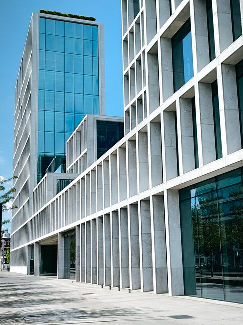 A modern multi-storey building with a predominantly reddish-brown exterior featuring vertical wooden slats and large glass windows. The building is situated on a paved pedestrian pathway bordered by a