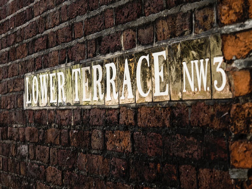 Close-up image of a street nameplate attached to a red brick wall. The sign reads 'Lower Terrace NW3' with white capital letters on a metallic background that shows signs of weathering and moss, positioned horizontally. The brick wall exhibits a mix of dark and reddish-brown hues with visible mortar lines. The scene is outdoors with natural lighting, and the focus is on the signage, suggesting a residential area typical of locations involved in home relocations or moving preparations, which relates to the services offered by Man with Van Denmark Hill within the removals sector.