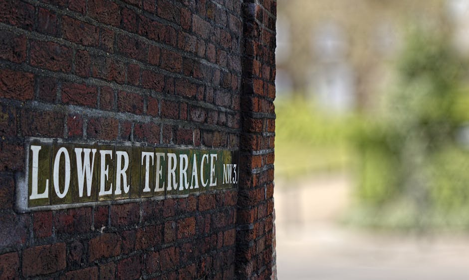 Close-up image of a street nameplate attached to a red brick wall. The sign reads 'Lower Terrace NW3' with white capital letters on a metallic background that shows signs of weathering and moss, positioned horizontally. The brick wall exhibits a mix of dark and reddish-brown hues with visible mortar lines. The scene is outdoors with natural lighting, and the focus is on the signage, suggesting a residential area typical of locations involved in home relocations or moving preparations, which relates to the services offered by Man with Van Denmark Hill within the removals sector.
