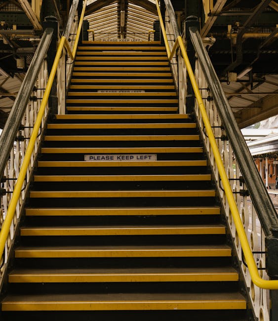 A set of industrial stairs leading up from an indoor loading area, with yellow safety handrails on both sides and black anti-slip strips on each step. The staircase is constructed of metal and wood, with a sign on the fourth step that reads 'PLEASE KEEP LEFT.' The environment appears to be a commercial or warehouse space, possibly part of a transportation or moving facility, with visible metal beams, scaffolding, and lighting fixtures overhead. This staircase is part of the logistics process involved in home relocation or furniture transport, supporting organized movement of goods between different levels of a building. The setting is well-lit, emphasizing the structural details of the staircase and surrounding environment, consistent with professional removals services such as those provided by Man with Van Denmark Hill.
