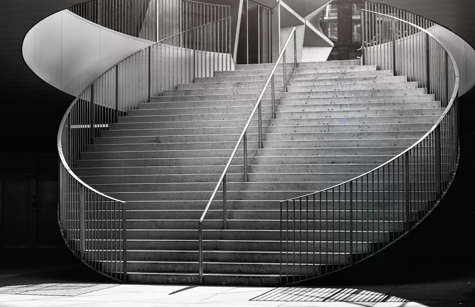 A black and white photograph of a large, curved concrete staircase with metal railings on both sides, located indoors beneath a modern, architectural ceiling structure. The staircase ascends from the floor to an upper level with wide steps and a central handrail. The surrounding environment appears to be a contemporary public space or building interior, with minimalistic design elements. The lighting creates shadows cast by the railings onto the steps, emphasizing the geometric shapes and the spiral form of the staircase. This setting is suitable for house removals or furniture transport processes involved in home relocation, as captured during the loading or unloading phase, possibly by Man with Van Denmark Hill, near Denmark Hill station.