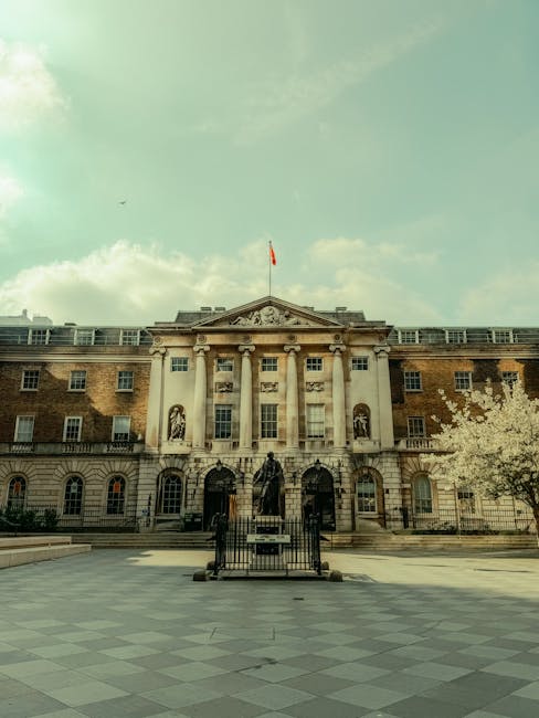 The image shows a historic multi-storey building with classical architectural features, including tall columns and decorative stonework, situated in an urban area with a paved open space in front. The building's facade combines brick and stone elements, and a flag is flying atop the central section. In the foreground, there is a statue or sculpture positioned on a pedestal, enclosed by a low metal fence, with a set of steps leading up to it. To the right, there is a tree with white blossoms. The scene is lit by natural daylight, with a partly cloudy sky overhead. This setting could relate to a university, government building, or civic institution, often involved in relocation or administrative operations. Man with Van Denmark Hill would use such a location to facilitate home relocation or furniture transport services, ensuring efficient access for moving equipment or vehicles involved in house removals near King's College Hospital.