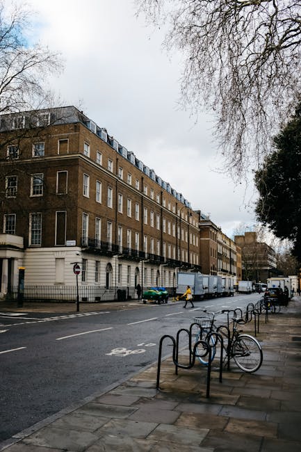 A wide street view in an urban residential area with a large, multi-storey brick building on the left side, featuring white window frames and balconies on the upper floors. The street has several parked bicycles secured to metal racks along the pavement, and a few pedestrians walking on the sidewalk. A blue van and a medium-sized green truck are parked in front of the building, with the truck positioned at an angle near the entrance, likely being loaded or unloaded during a home relocation process. The scene is illuminated by natural daylight on an overcast day, with leafless trees visible at the top right and background. The surroundings include other residential buildings and parked vehicles, reflecting typical moving logistics in a city environment, with Man with Van Denmark Hill potentially involved in furniture transport or packing and moving services near Denmark Hill and King's College Hospital.