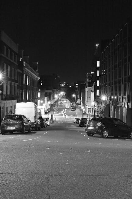 Black and white nighttime photograph capturing a city street scene with parked cars along the curbside, including a small van and several passenger vehicles, positioned near residential and commercial