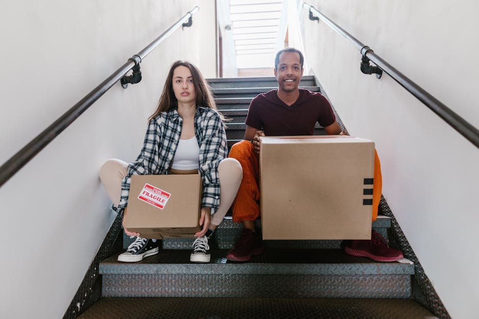 A man and a woman sitting on indoor staircase steps during a home relocation, each holding cardboard boxes for furniture transport and packing and moving purposes. The woman, seated on the left, has long brown hair, and is wearing a black-and-white plaid shirt, white top, and black-and-white sneakers. She holds a medium-sized, sealed cardboard box labeled 'fragile.' The man, on the right, has short dark hair, a light beard, and is dressed in a maroon t-shirt and orange pants. He holds a larger cardboard box with black stripes. The staircase has black anti-slip treads, a white wall on both sides, and metal handrails attached to the walls. Behind them, a doorway with a partly open door leads to a well-lit room with wooden flooring, suggesting they are preparing for or in the process of a house move, with visible materials like packing tape and cushions. This scene exemplifies the process of furniture and belongings being carefully loaded for a home relocation, coordinated by a professional removal service, such as Man with Van Denmark Hill, in an urban residential setting.