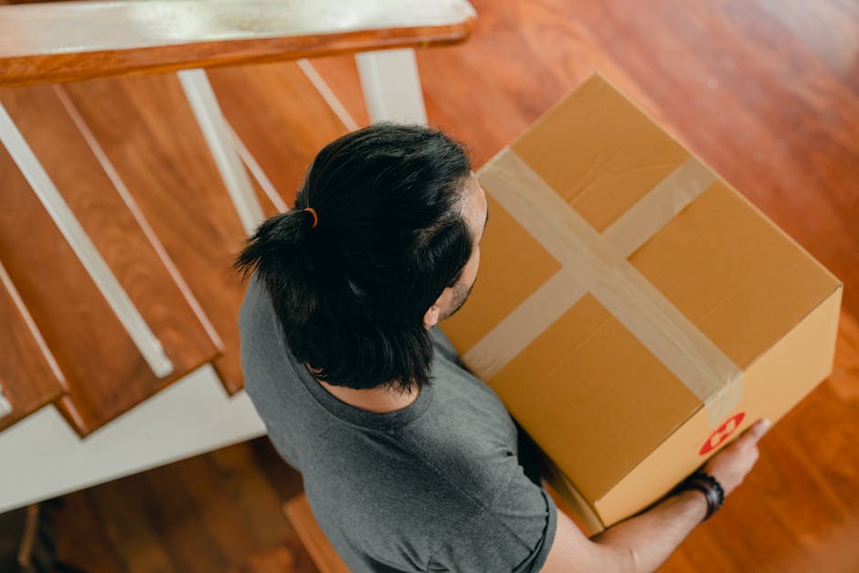 A man with long dark hair tied back in a ponytail, wearing a grey T-shirt and wristwatch, is holding a large cardboard box sealed with tan packing tape, featuring a red recycling symbol on the side. He is positioned indoors on a wooden staircase with white risers and a wooden banister, near a doorway, preparing to move or load the box. The environment shows a well-lit interior space with natural light, and the scene captures the process of packing or furniture transport as part of a home relocation service by Man with Van Denmark Hill, focusing on moving logistics suited for properties with narrow staircases.