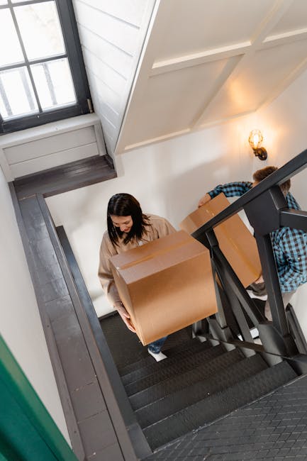 A man and a woman sitting on indoor staircase steps during a home relocation, each holding cardboard boxes for furniture transport and packing and moving purposes. The woman, seated on the left, has long brown hair, and is wearing a black-and-white plaid shirt, white top, and black-and-white sneakers. She holds a medium-sized, sealed cardboard box labeled 'fragile.' The man, on the right, has short dark hair, a light beard, and is dressed in a maroon t-shirt and orange pants. He holds a larger cardboard box with black stripes. The staircase has black anti-slip treads, a white wall on both sides, and metal handrails attached to the walls. Behind them, a doorway with a partly open door leads to a well-lit room with wooden flooring, suggesting they are preparing for or in the process of a house move, with visible materials like packing tape and cushions. This scene exemplifies the process of furniture and belongings being carefully loaded for a home relocation, coordinated by a professional removal service, such as Man with Van Denmark Hill, in an urban residential setting.