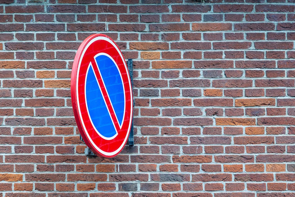 A round, red-bordered no parking sign with a blue background and a red diagonal line, mounted on a metal bracket attached to a red brick wall. The wall features horizontal rows of bricks in various shades of reddish-brown, with light gray mortar between them. The sign is positioned slightly to the left of the center of the image, with the wall extending evenly across the frame. Natural lighting illuminates the scene, highlighting the texture of the bricks and the reflective surface of the sign. This image relates to parking restrictions, which are relevant for home relocations and moving services in Southwark, as managed by Man with Van Denmark Hill, particularly when dealing with parking suspensions or fines associated with house removals.