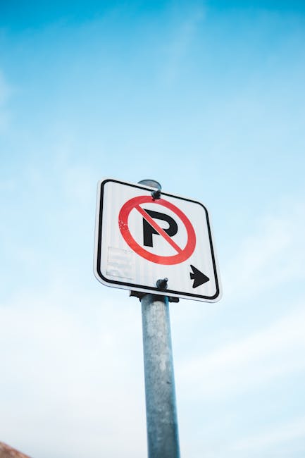 A metal parking restriction sign mounted on a grey pole against a partly cloudy sky, indicating no parking with a red circle and slash over a black 'P' symbol, and a black arrow pointing to the right; the sign is positioned near a building or street area in Southwark, relevant to house removals and moving logistics at Denmark Hill, with no vehicles or furniture visible in the image.