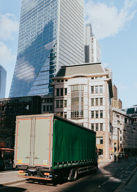 A large green and beige delivery truck parked on a city street with tall modern glass skyscrapers and older stone buildings in the background. The truck is positioned close to the curb, with its rear