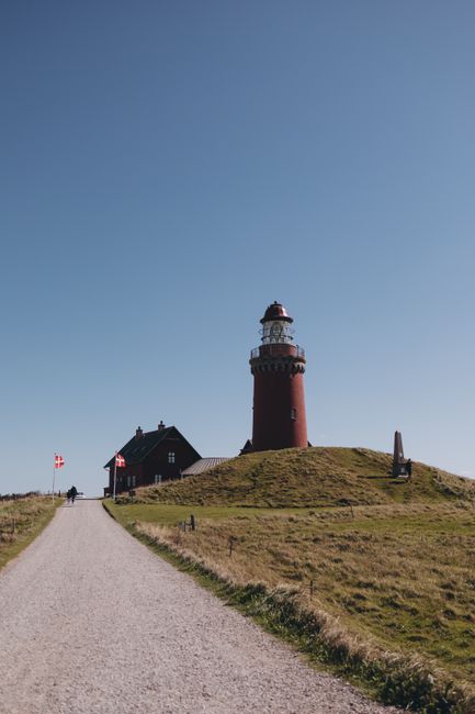 A tall, cylindrical red and white lighthouse situated atop a grassy hill, with a narrow gravel pathway leading up to it. The lighthouse has a black and white balcony near the top and a small dome-shaped lantern room with a black cover. Adjacent to the lighthouse, there is a small dark-colored building with a pitched roof, likely associated with the lighthouse station. The scene is set on a clear, sunny day with a bright blue sky and no clouds visible. In the foreground, the gravel path is bordered by short, green grass, and the landscape appears open and lightly hilly. The lighthouse and surrounding structures are part of an outdoor setting, with some distant figures visible near the base of the building, emphasizing the scale and prominence of the lighthouse within the landscape. This image may be used in the context of house removals or moving logistics related to relocating equipment or guiding transportation to coastal landmarks, with Man with Van Denmark Hill providing expert packing and moving services in similar settings.