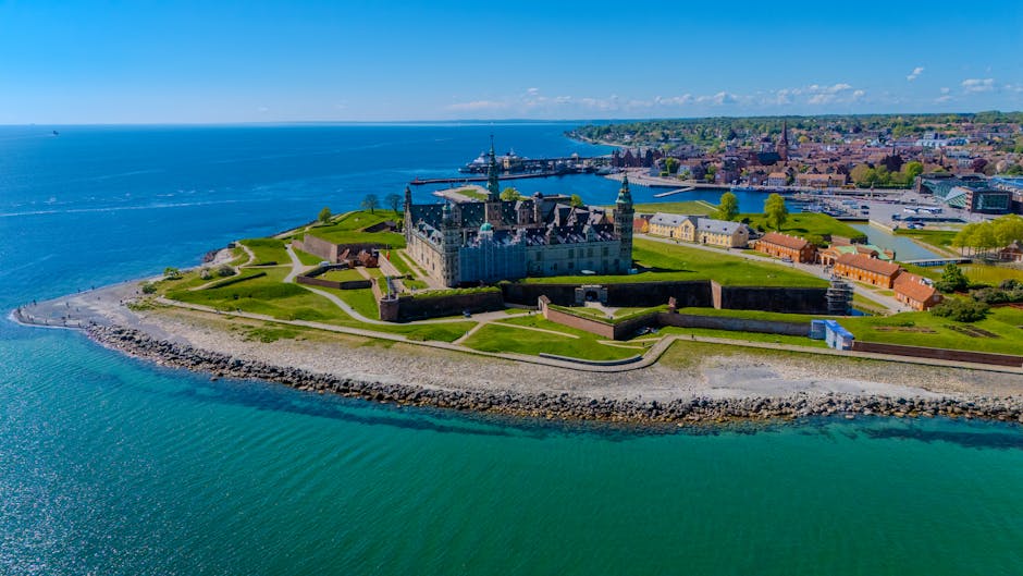 An aerial view of a historic castle situated on a small, fortified island surrounded by a broad expanse of water, with a coastal town and modern buildings visible in the background. The castle features a central structure with multiple turrets, stone walls, and a surrounding moat, all integrated into landscaped green lawns with pathways. The shoreline is protected by a stone breakwater, and the scene is captured in bright daylight under a clear sky, highlighting the contrast between the ancient architecture and the natural waterfront setting. This image showcases the coastal environment typical of house removals and relocation services in Denmark Hill, emphasizing the importance of careful planning during furniture transport and home relocation processes. Occasionally, Man with Van Denmark Hill manages the logistics of moving furniture and belongings in scenic, historic settings like this, ensuring efficient and secure transport despite challenging environments.
